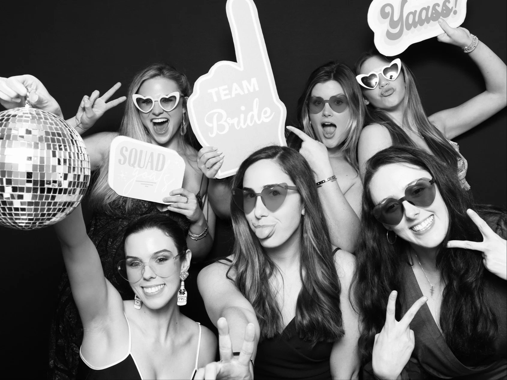 Group of six women at bridal party photo booth, holding signs that say "TEAM Bride," "Yaas!," and "Squad goals," wearing sunglasses, making peace signs, and having fun with a disco ball and playful expressions.