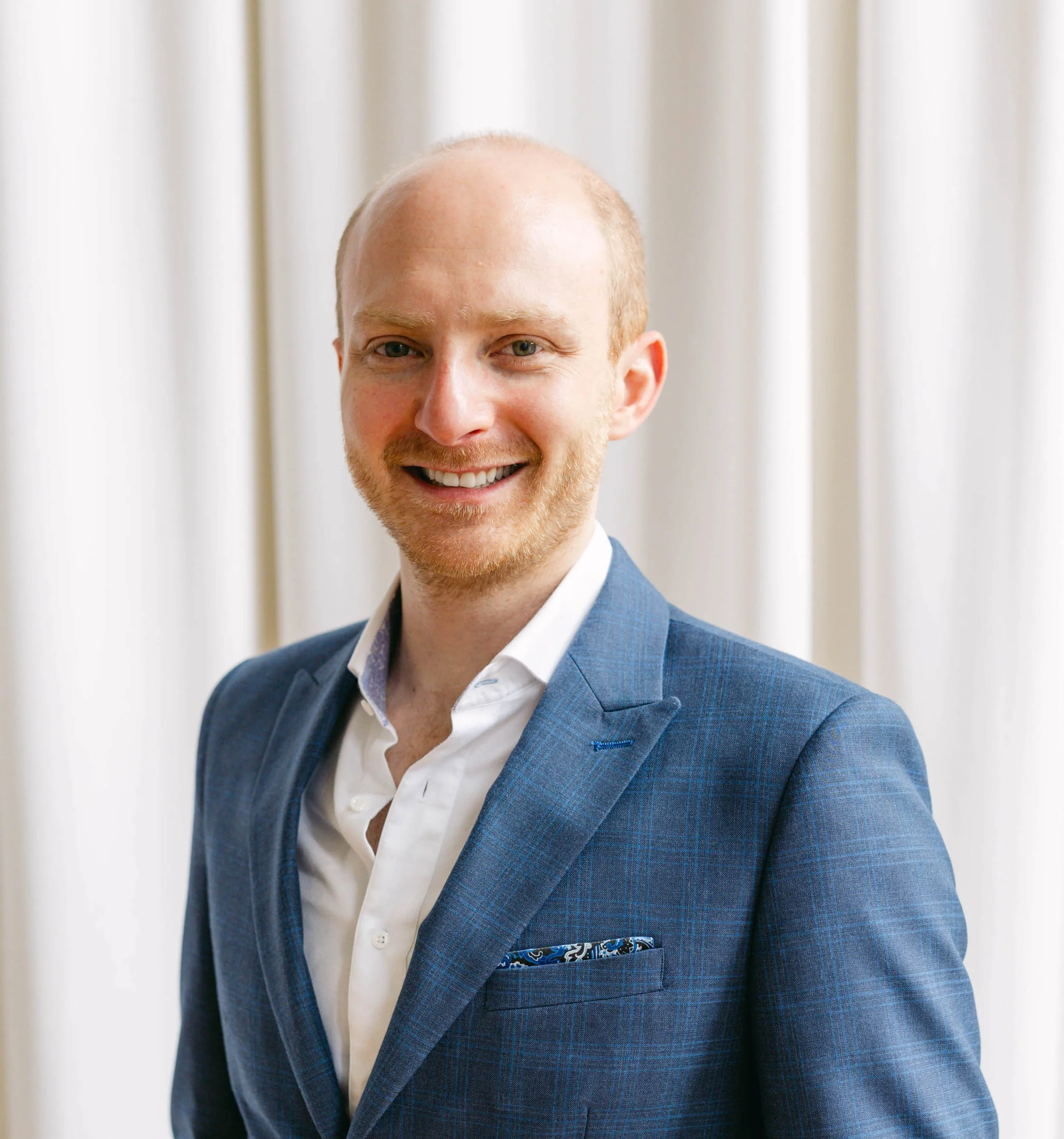 A man (Shane Pokroy, RP) smiling with a suit and a white shirt, standing in front of a light-colored curtain.