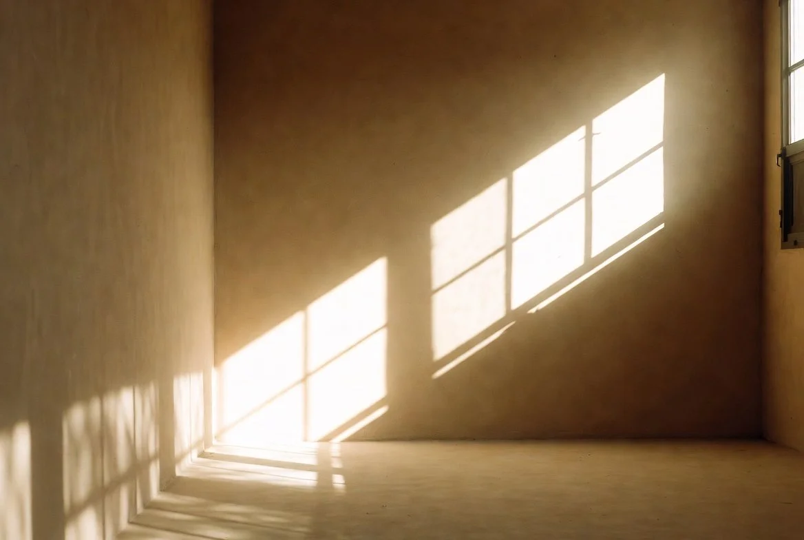 Sunlight streaming through a window casting shadowed grid patterns on an empty beige wall and floor.