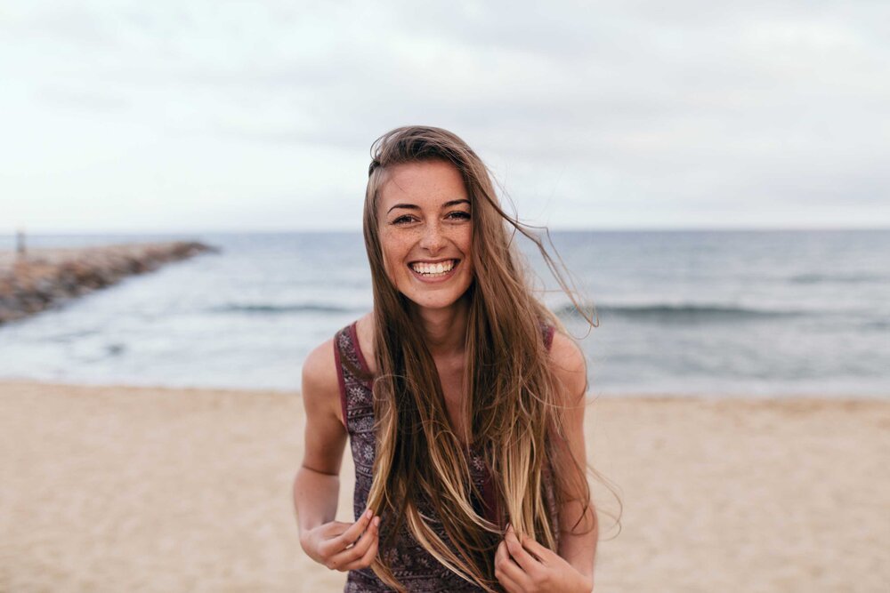 A young woman with long, wavy hair smiling at the camera on a beach, with the ocean and cloudy sky in the background.
