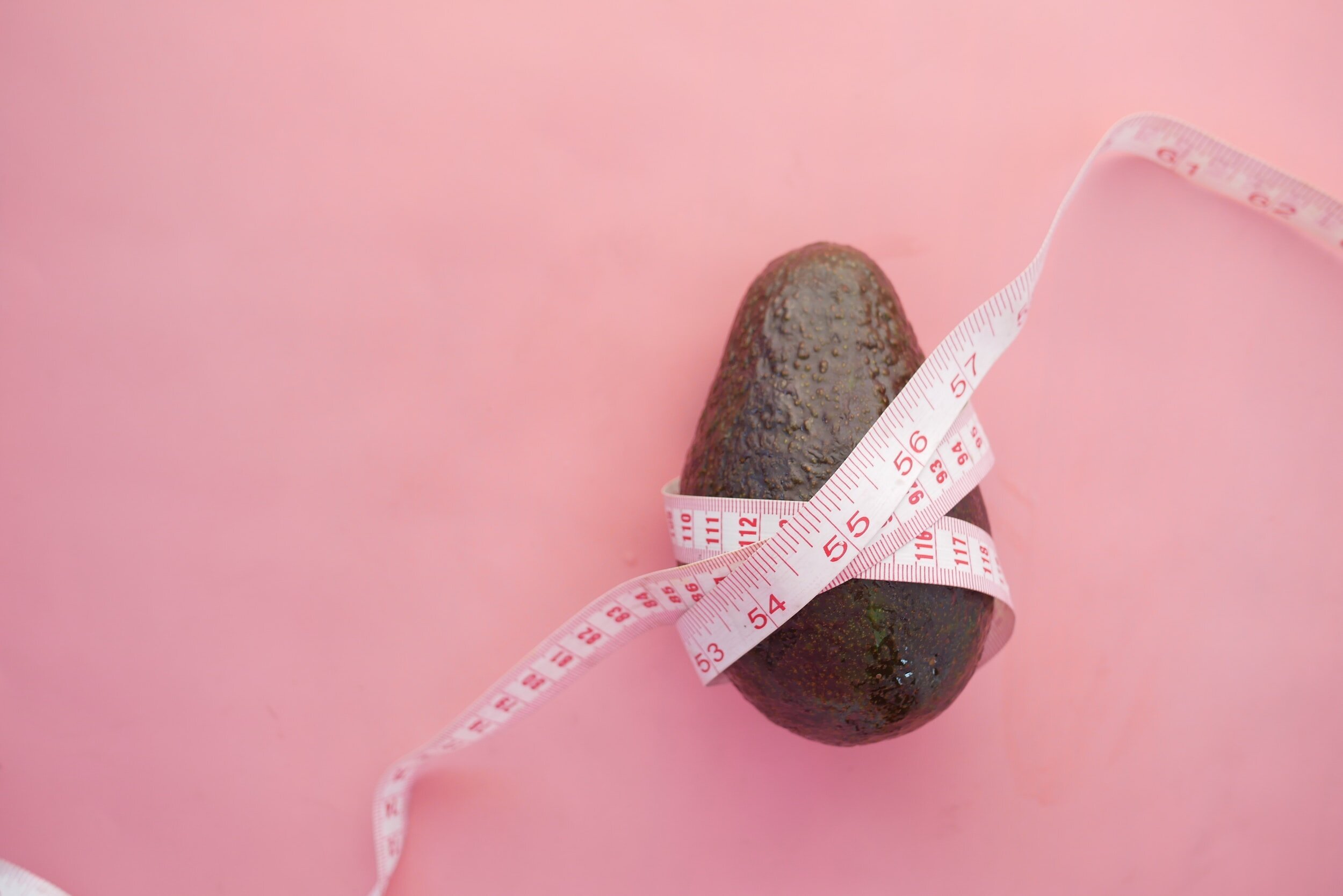 An avocado with a measuring tape wrapped around it placed on a pink background.