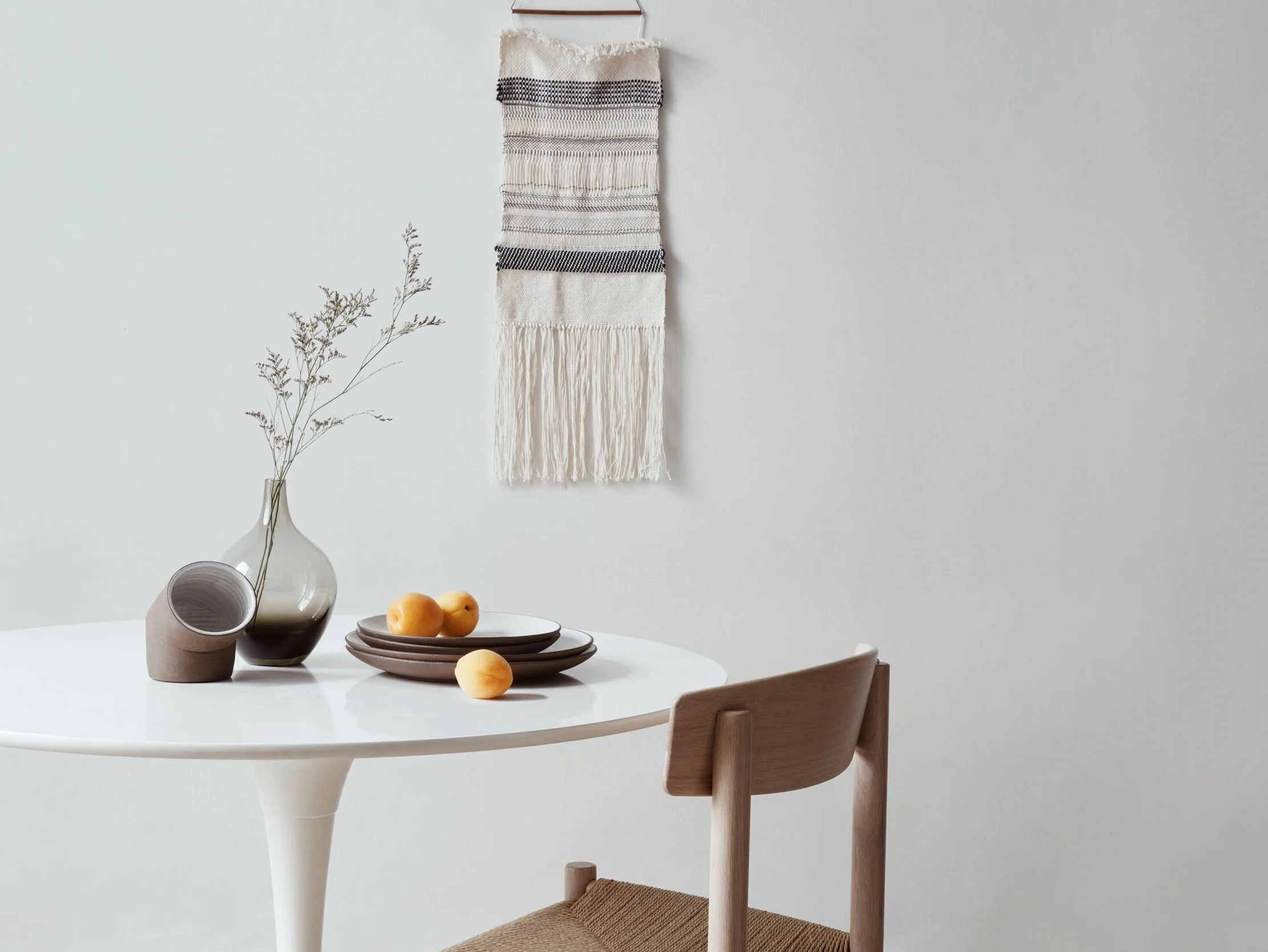 Minimalist dining area with a white round table, wooden chair, a beige vase with dried branches, a grey cup, grey plates with peaches, and a wall hanging woven textile with fringe, all set against a plain white wall.