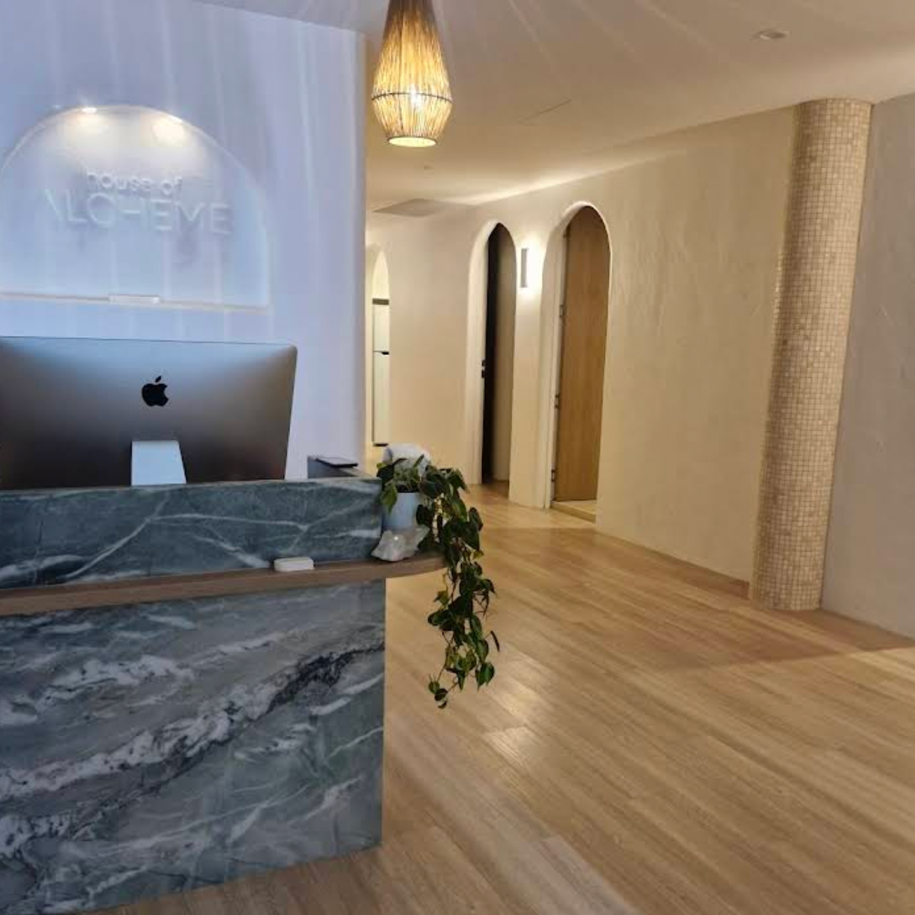 Interior of a modern reception area with wooden floors, a marble counter with a plant, and arched doorways in the background.