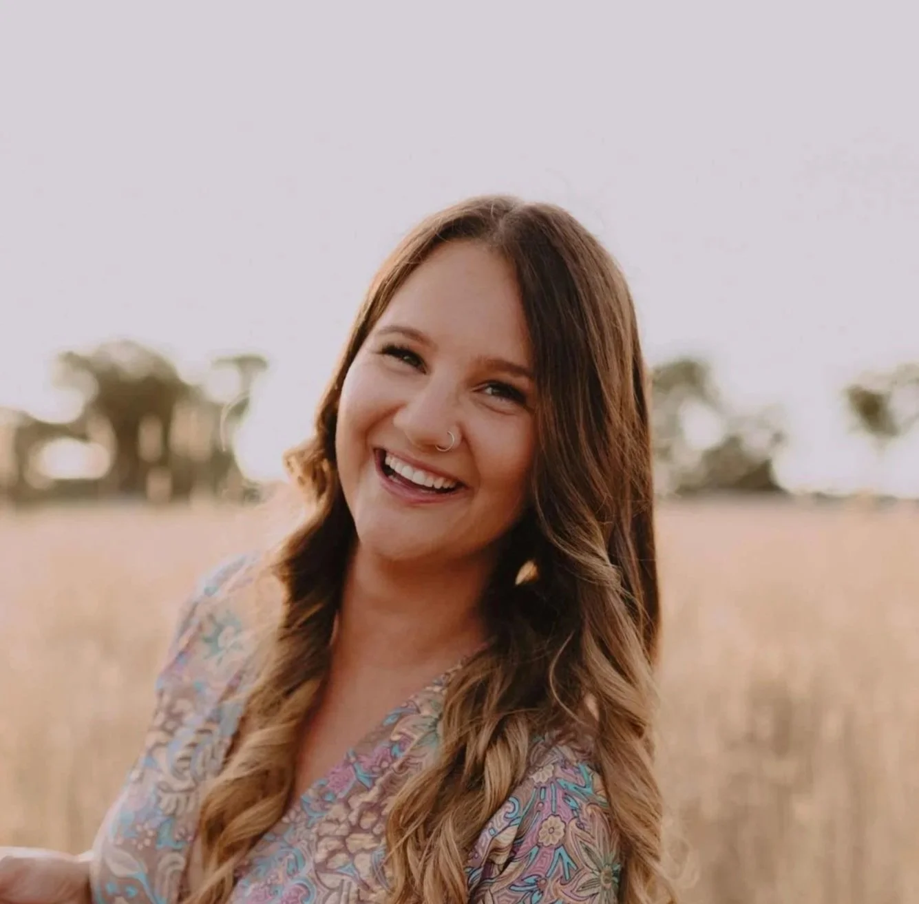 A young woman with long, wavy brown hair, smiling and looking towards the camera outdoors in a field with trees in the distance, during the daytime.