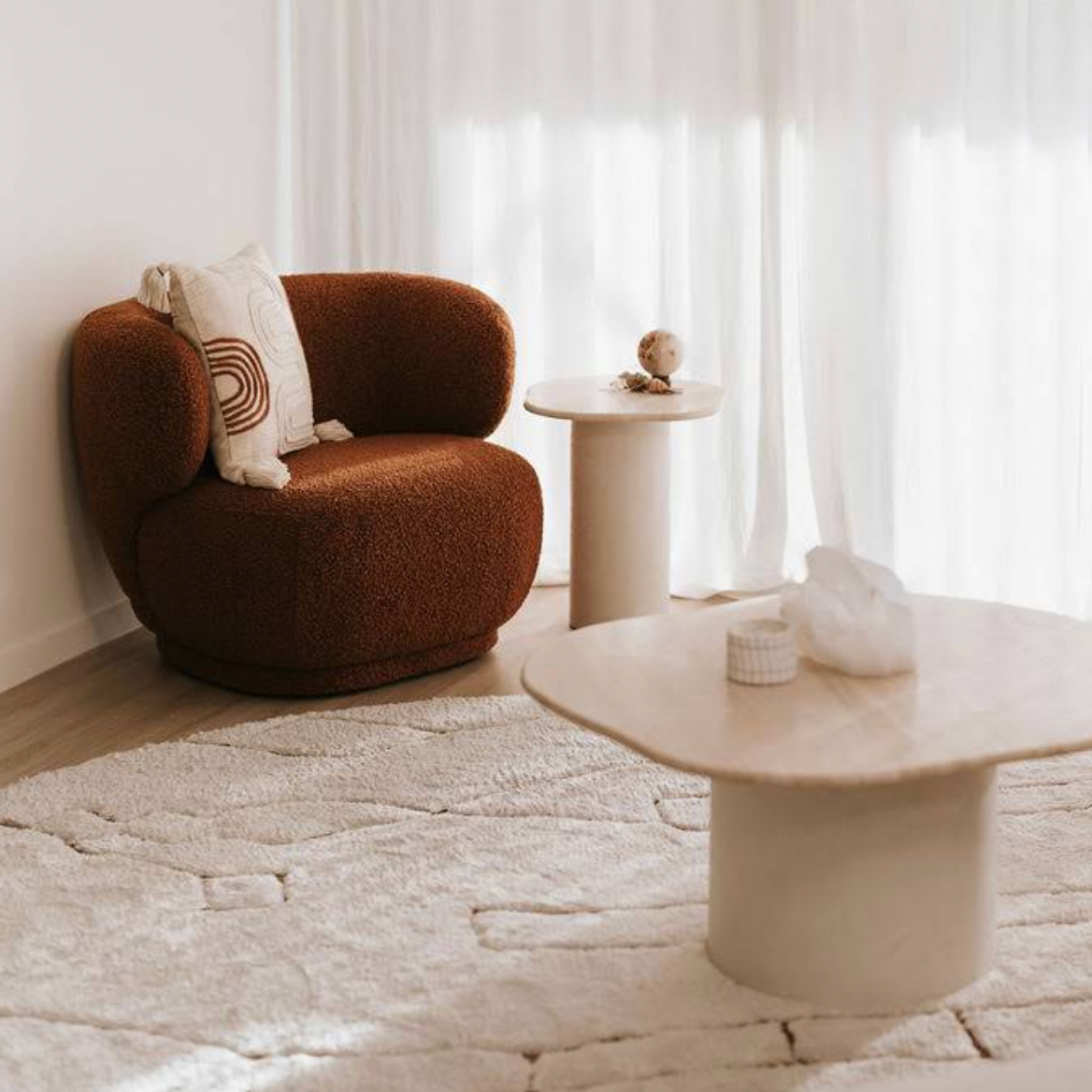 Living room with a rust-colored textured armchair, white decorative pillows, a small round white side table with a globe and decorative items, a larger white table with objects, a plush cream rug, and sheer white curtains letting in natural light.