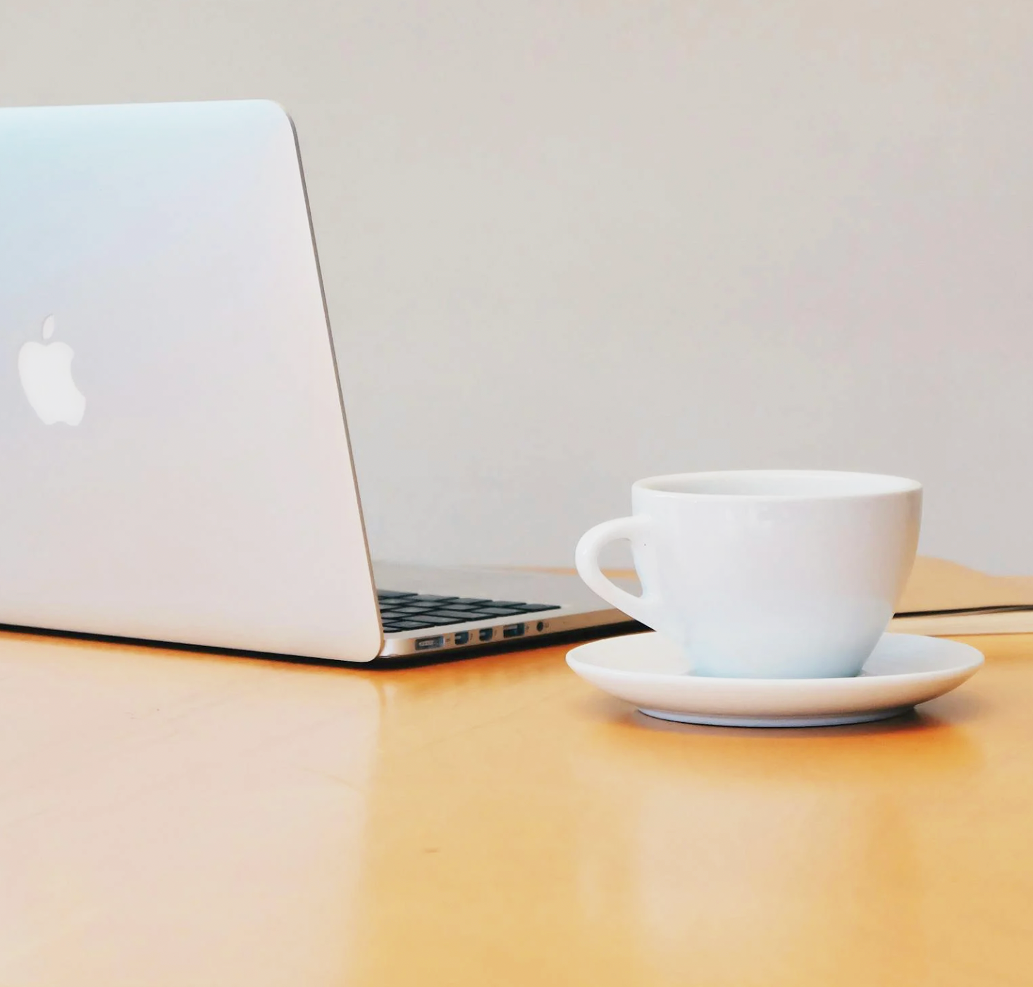 Close-up of a MacBook laptop and a white coffee cup on a saucer on a wooden desk.