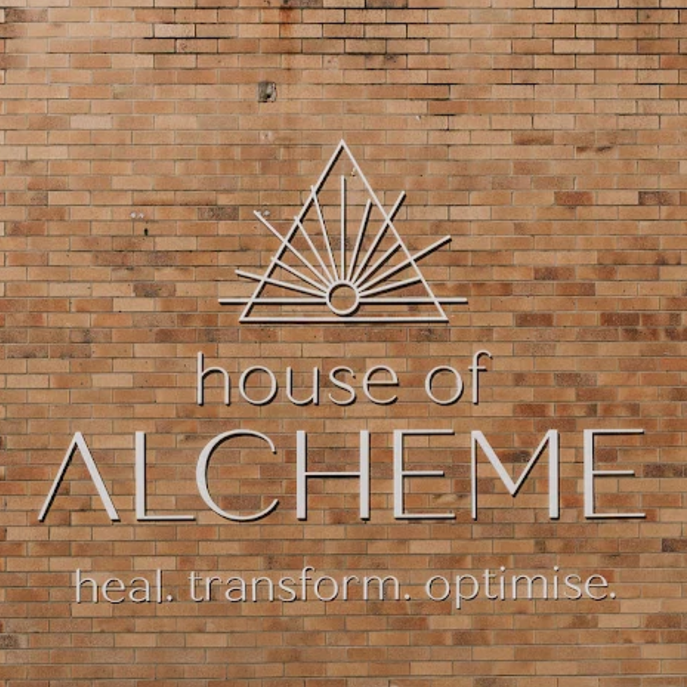 Sign on a brick wall for house of ALchemy with a geometric design resembling a house or pyramid and sun rays, and the words 'heal. transform. optimise.' underneath.