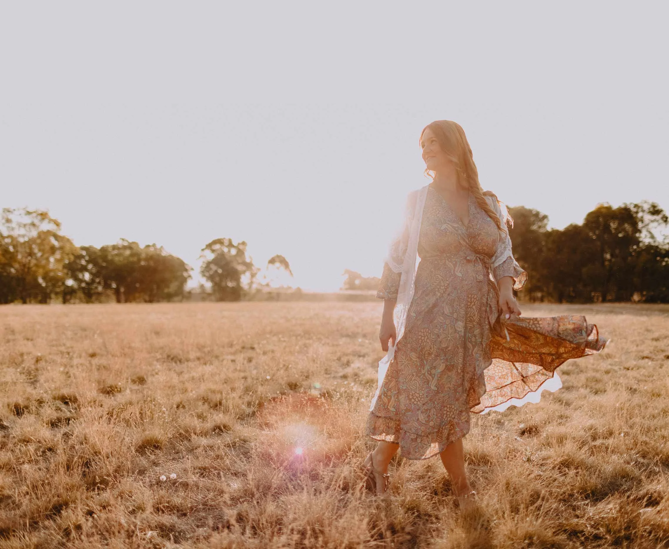A woman with long hair wearing a floral dress walking through a field at sunset, smiling and looking to the side.