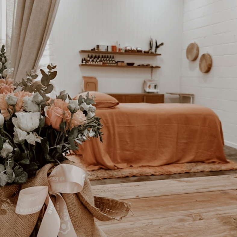 A cozy bedroom featuring a bed with an orange bedspread and matching pillow, a wooden headboard, a white wall with two wicker wall decorations, a floating shelf with various items, and a bouquet of flowers in the foreground wrapped in burlap with a white ribbon.