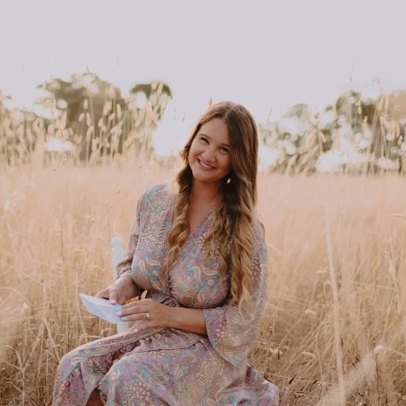 Young woman sitting in a field of tall grass, smiling and holding a piece of paper.