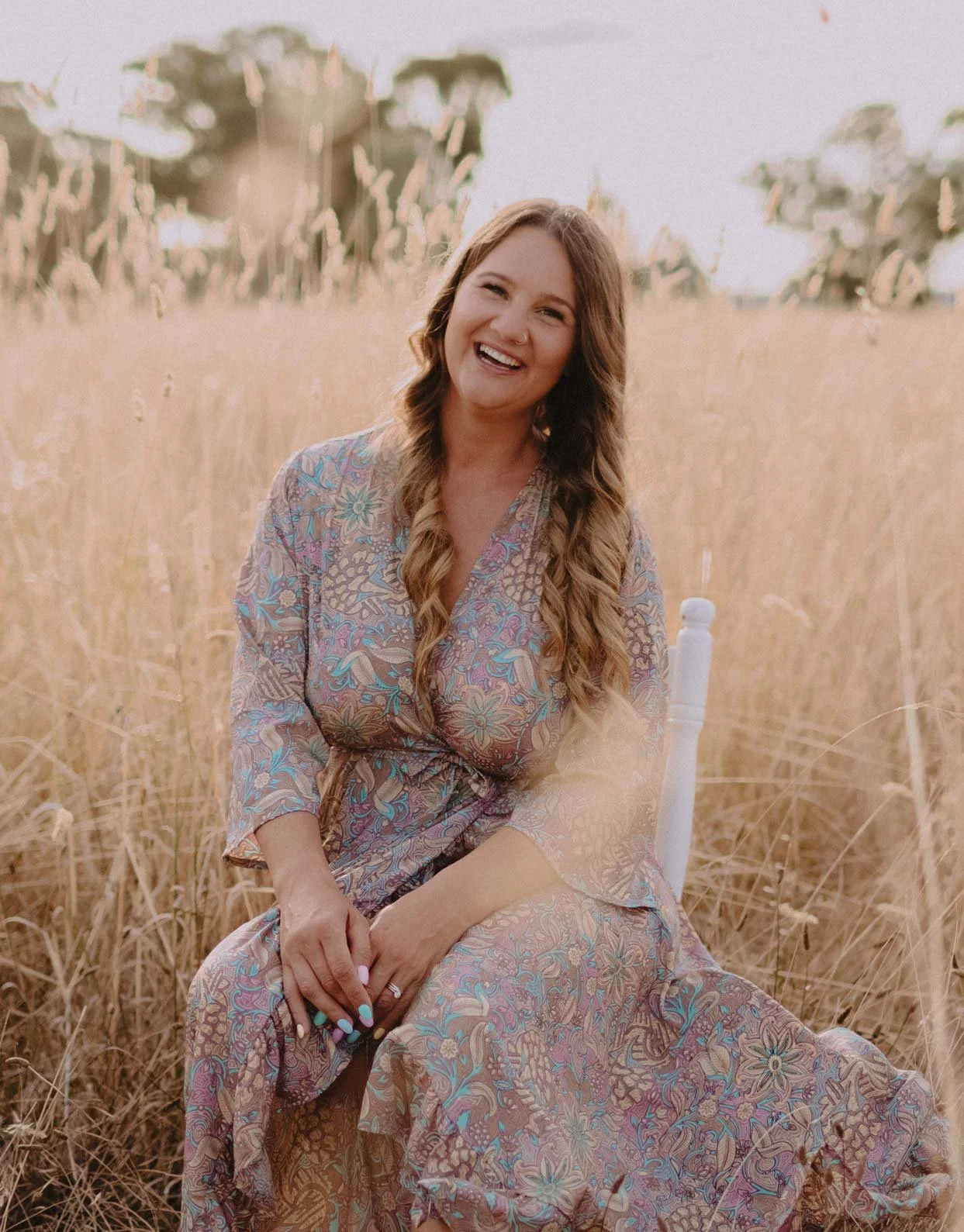 A woman with long wavy hair sitting in a field of tall dry grass, smiling and wearing a patterned dress.