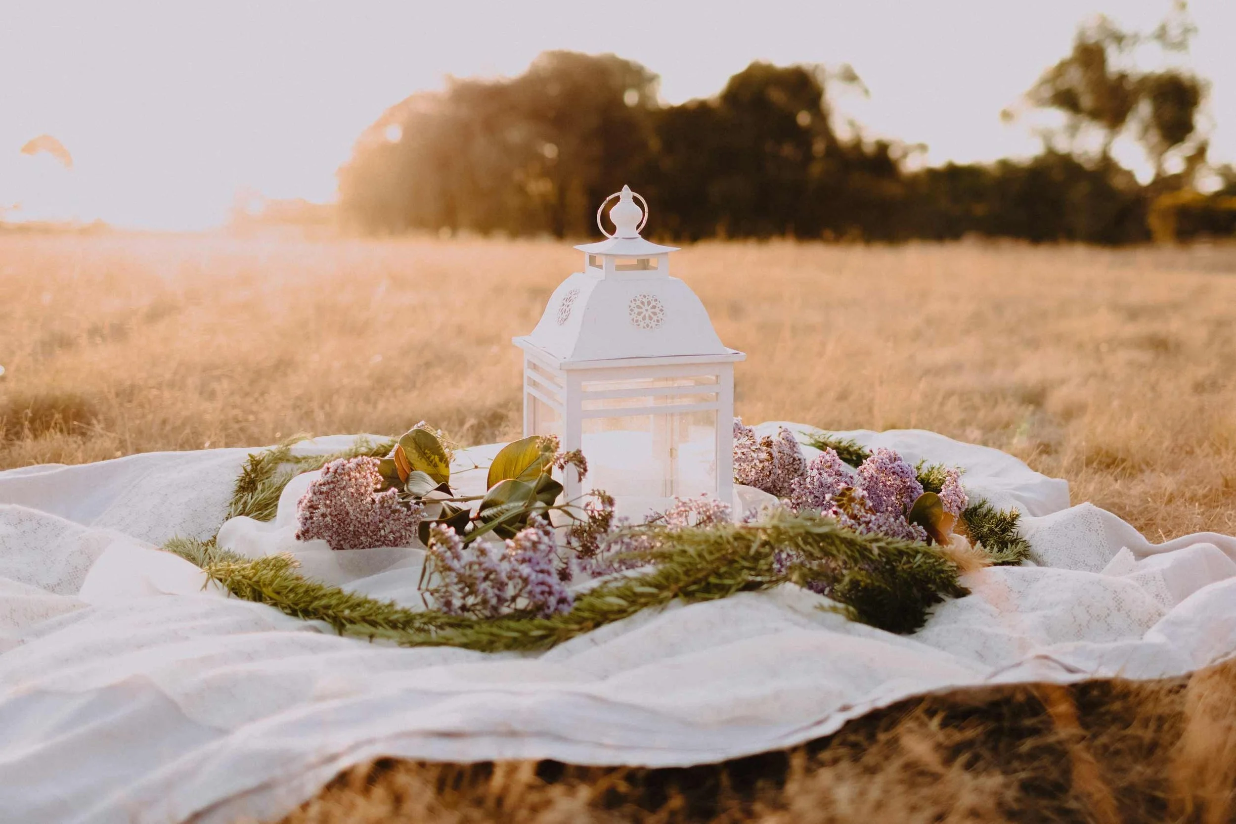A white lantern surrounded by pink and green flowers rests on white fabric in an outdoor setting during sunset or sunrise.