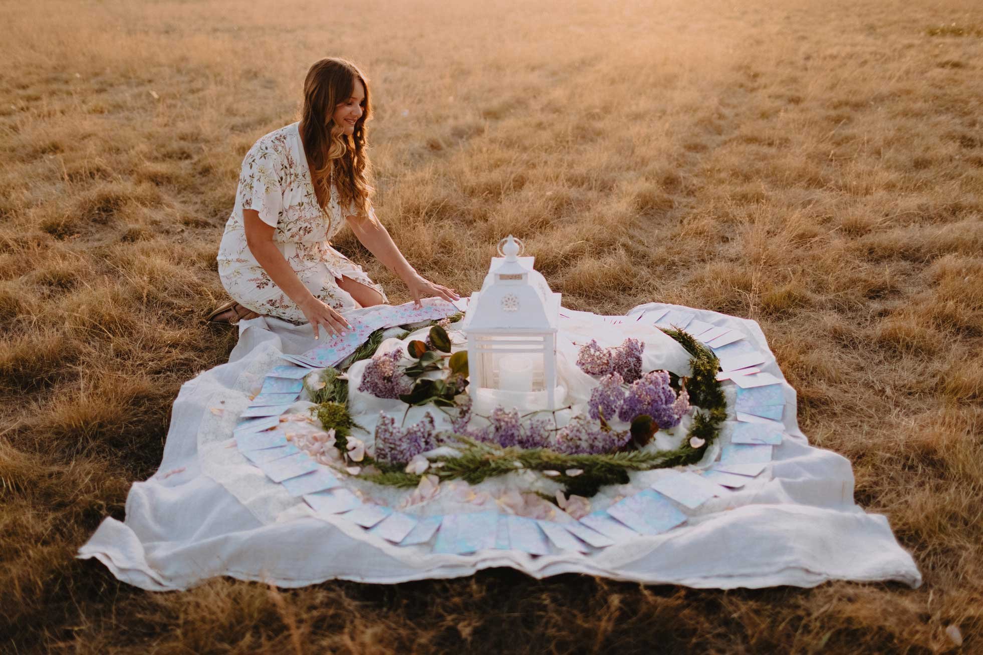 A woman in a floral dress kneels on a white blanket spread on dry grass in a field, arranging a floral display and lanterns for an outdoor celebration at sunset.