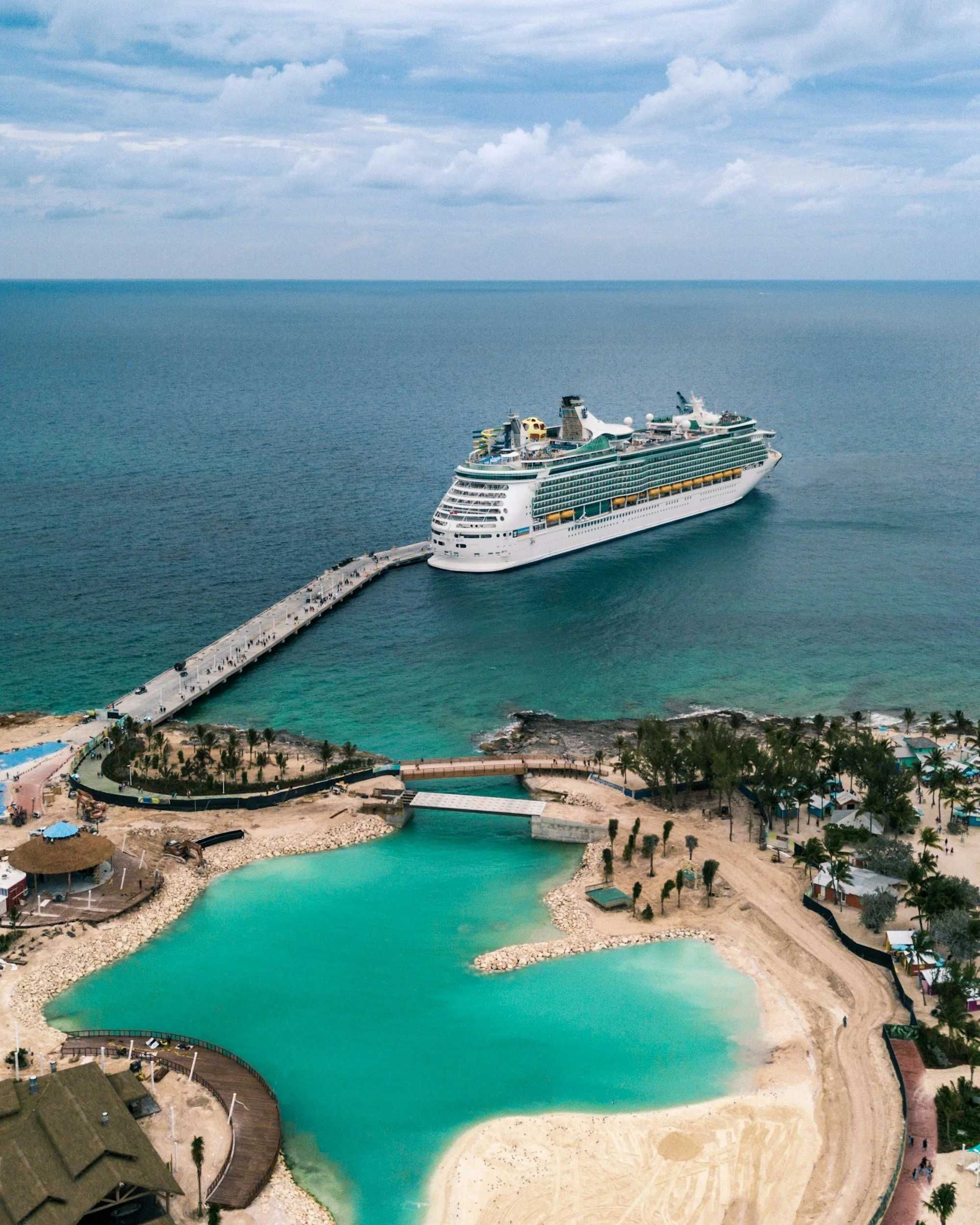 A cruise ship docked at a pier extending into a body of water, with a beach and some buildings in the foreground and a cloudy sky overhead.