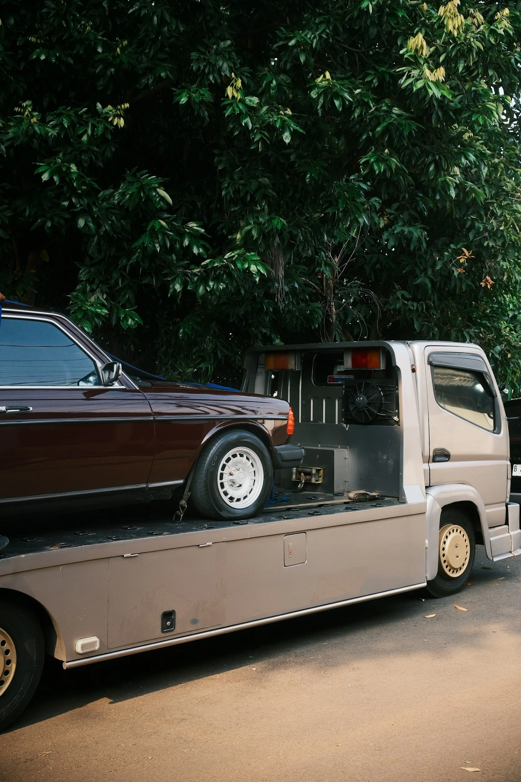 A beige flatbed tow truck carrying a vintage dark red car with white wheels parked on a street, with a large green leafy tree in the background.