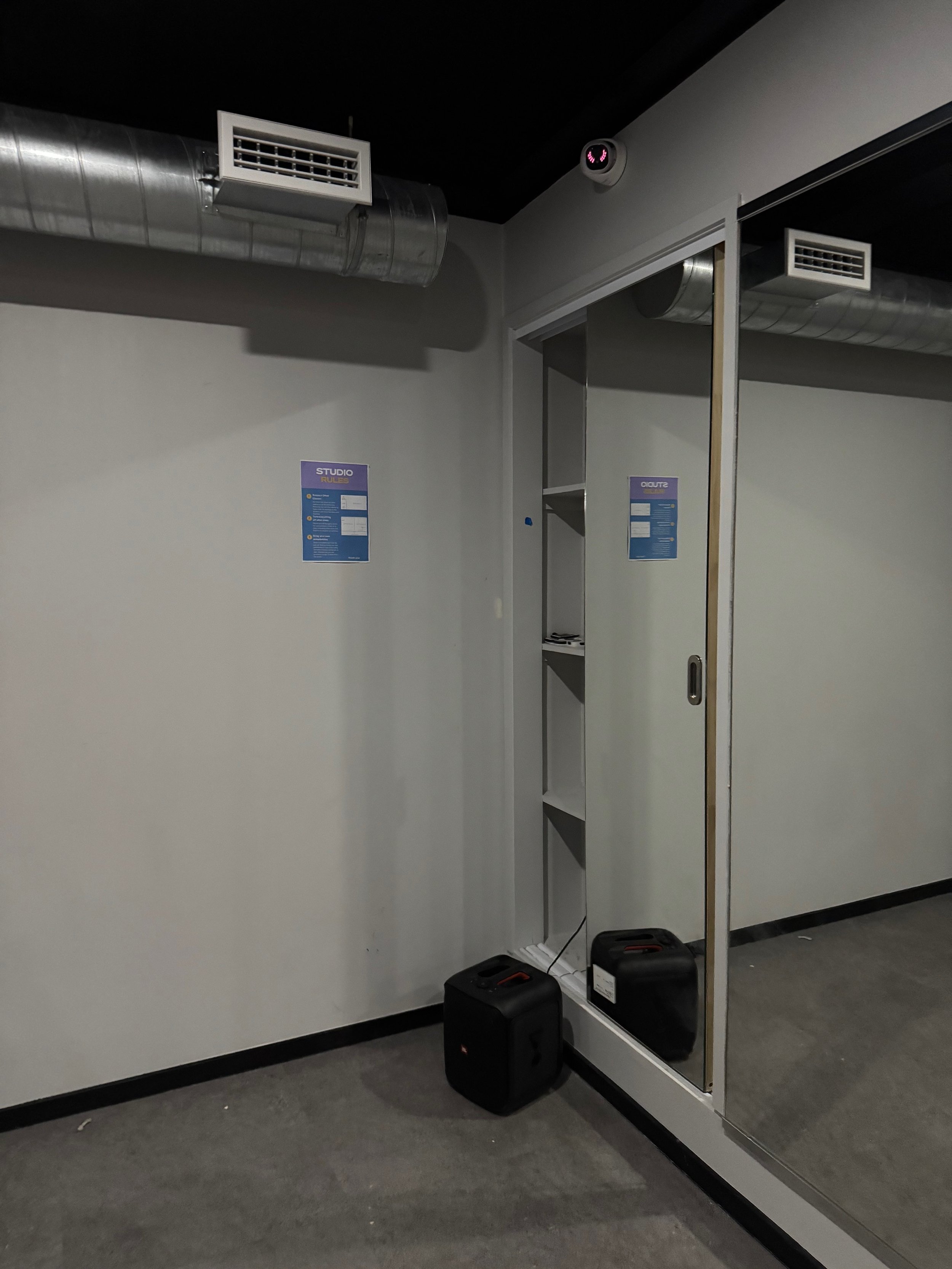 A room corner with an air duct, security camera, mirror, and speaker on a carpeted floor.