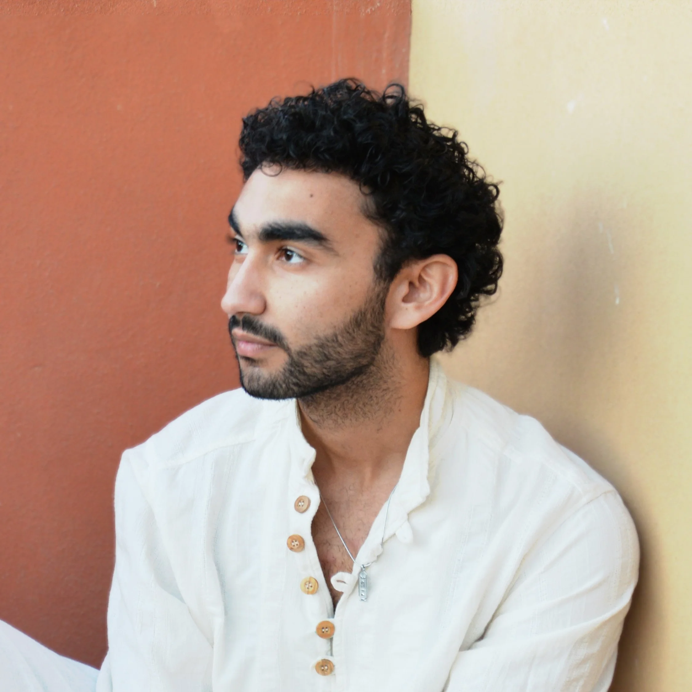 Profile of a young man with curly black hair, tan skin, and a beard, wearing a cream-colored shirt and a necklace, sitting against a wall with warm tones.