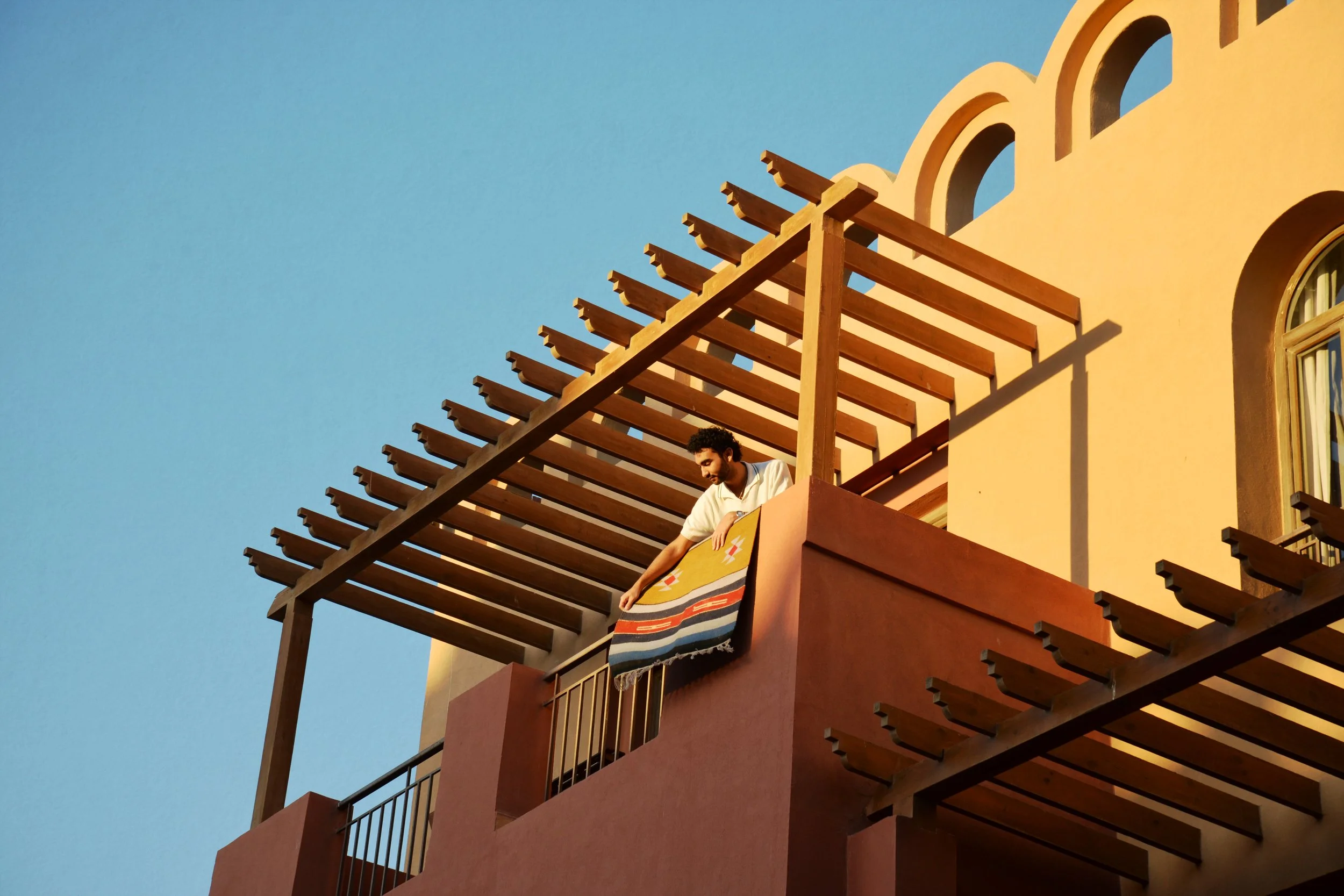 A man with curly hair and a beard holds a colorful striped towel while standing on the balcony of a yellow building with wooden beams against a clear blue sky.