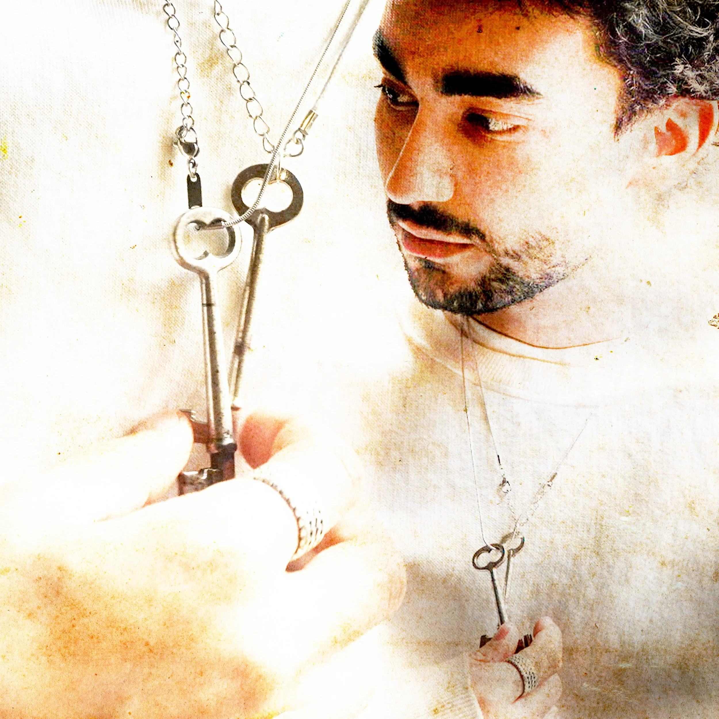 A man with dark hair and a beard is holding two vintage keys on a necklace close to his face, looking at them with interest.