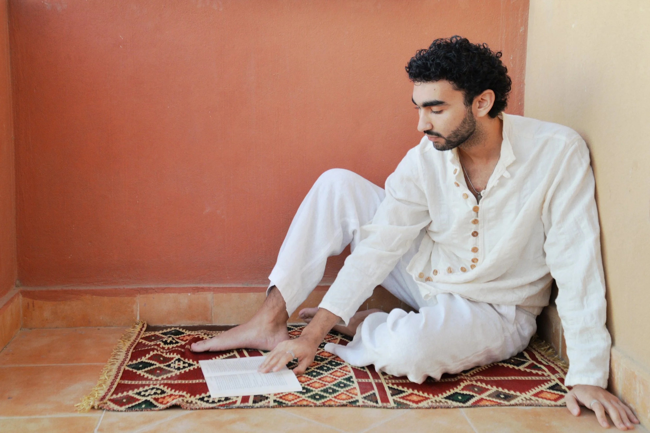 A man with curly dark hair, wearing a beige traditional outfit, sitting cross-legged on a patterned rug on the floor, reading a book, against a two-tone orange and yellow wall.