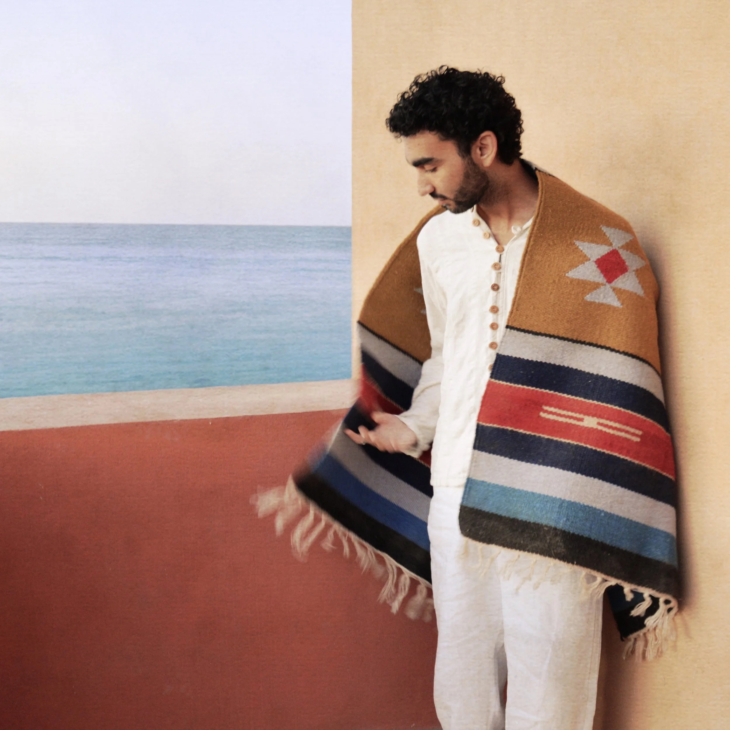 A man standing indoors against a beige wall, wearing a colorful woven shawl, white shirt, and pants, looking down while holding the shawl.