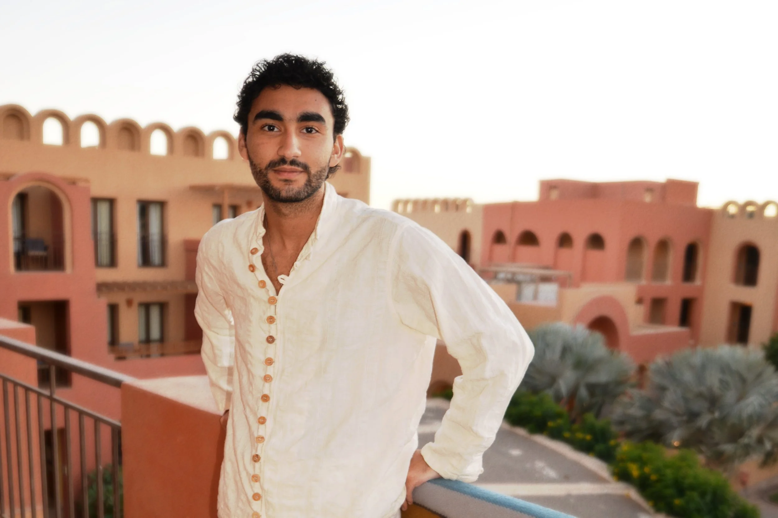 A man with dark curly hair and a beard standing on a balcony, wearing a cream-colored shirt with wooden buttons, with a peach-colored building with arched windows and lush greenery in the background during sunset.