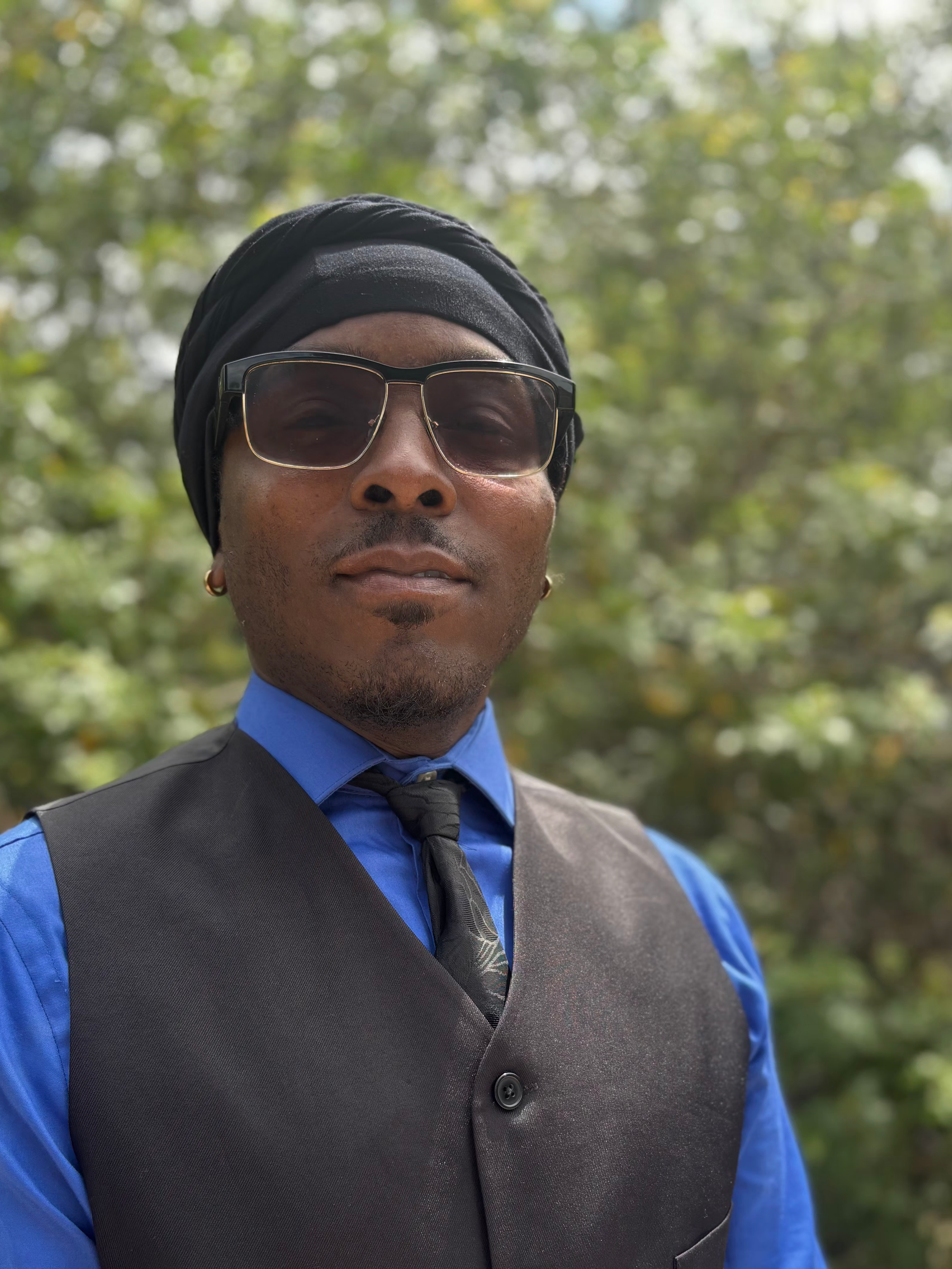 Close-up of a man wearing sunglasses, a black headwrap, a blue shirt, a black vest, and a gray tie, outdoors with green trees in the background.