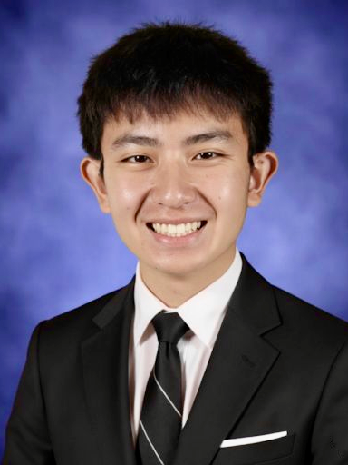 Young man in a black suit with a white shirt and black tie, smiling in front of a blue background.