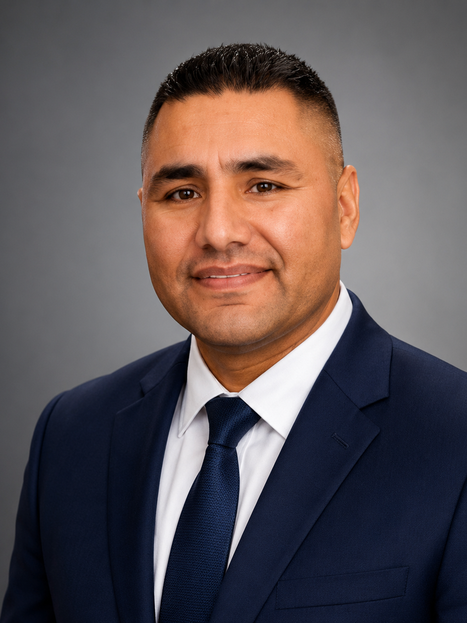 Professional headshot of a man in a dark blue suit, white shirt, and dark blue tie, smiling against a gray background.