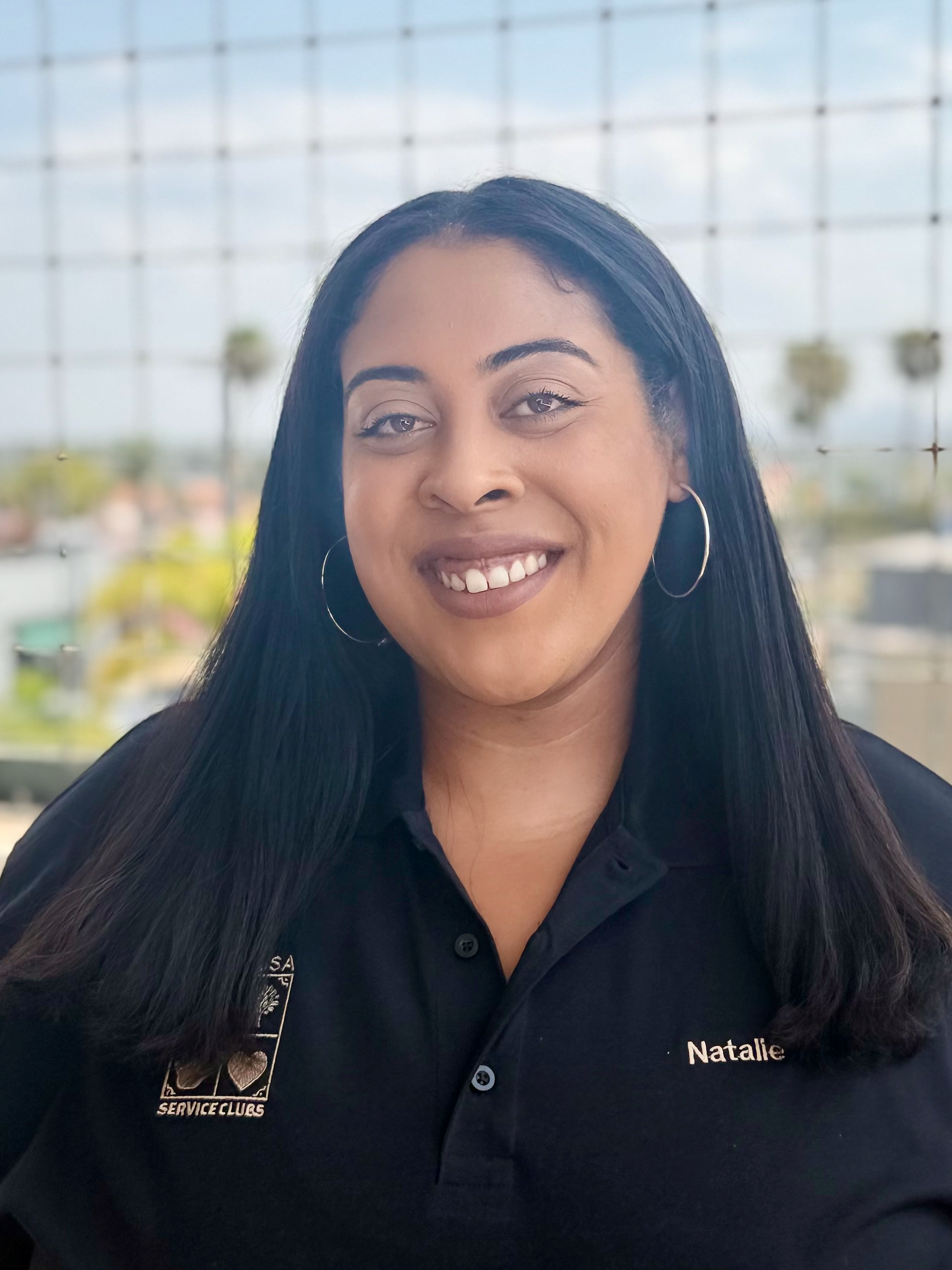 Close-up of a smiling woman with long black hair wearing a black polo shirt with name tag 'Natalie' and patches, standing in front of a blurred background of a building with hanging plant baskets.