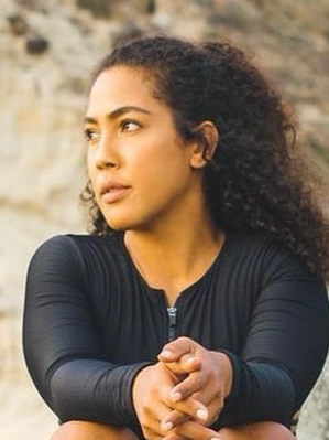 A woman with curly hair sitting outdoors in front of a rocky background, wearing a black long-sleeve top.