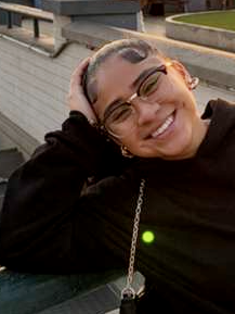 A woman smiling and relaxing outdoors, wearing glasses and a black top, with her hand behind her head, near a body of water and a brick wall.