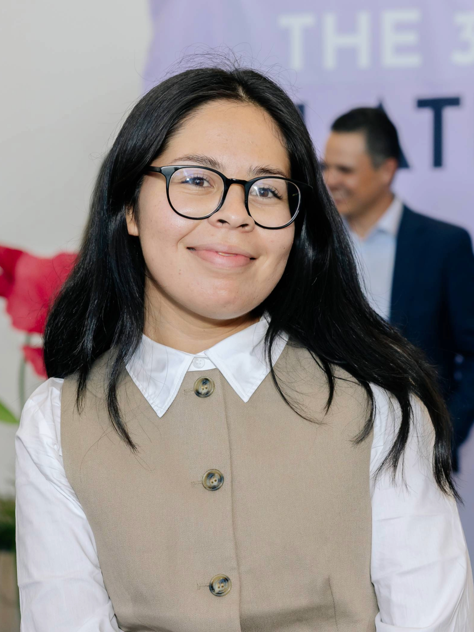 Close-up of a young woman with long black hair, wearing glasses, a white collared shirt, and a beige vest, smiling at the camera. In the background, a man in a suit and a blurred purple sign with partially visible white text.