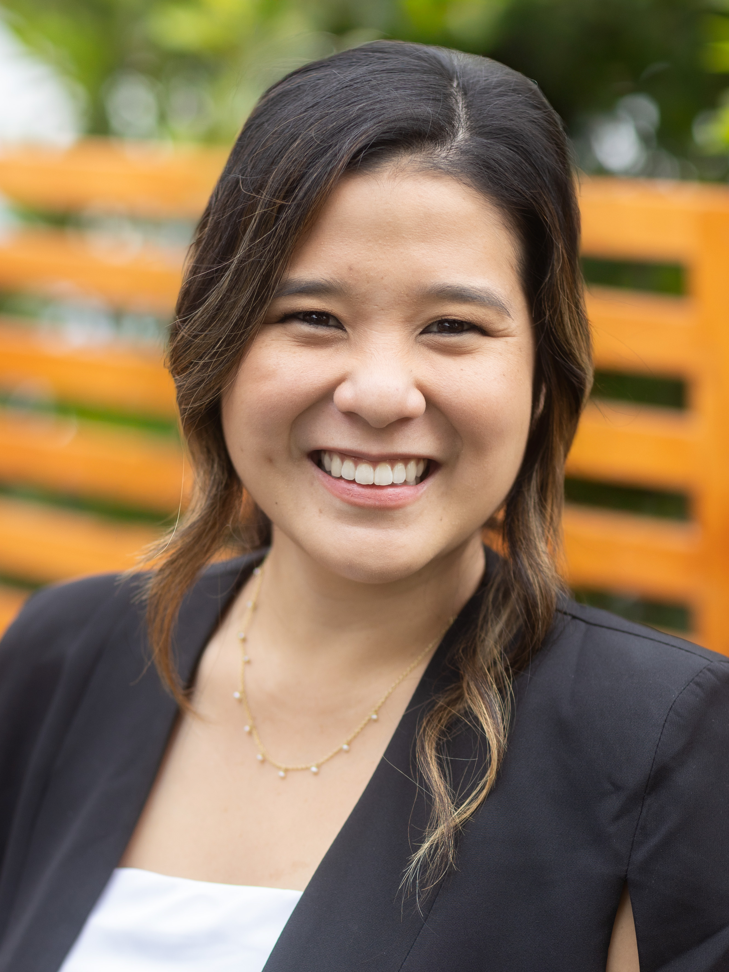 Close-up of a woman with dark hair smiling outdoors, wearing a black blazer and a gold necklace, with a blurred wooden fence and green foliage in the background.