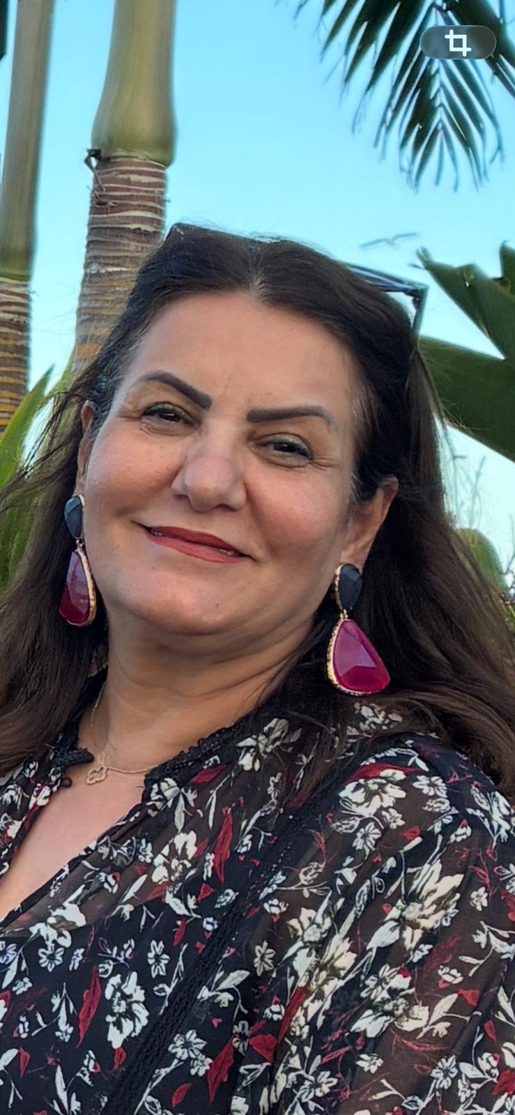 A woman smiling outdoors with palm trees and a clear blue sky in the background.
