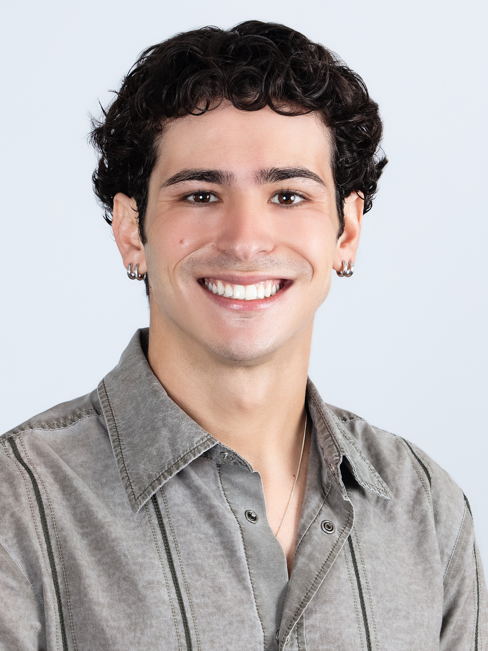 Close-up of a smiling person with short, curly dark hair, earrings, wearing a grey button-up shirt with dark stitching, against a plain white background.
