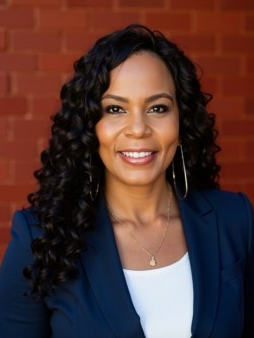 Portrait of a woman with curly black hair, wearing a navy blazer and white top, smiling in front of a brick wall.