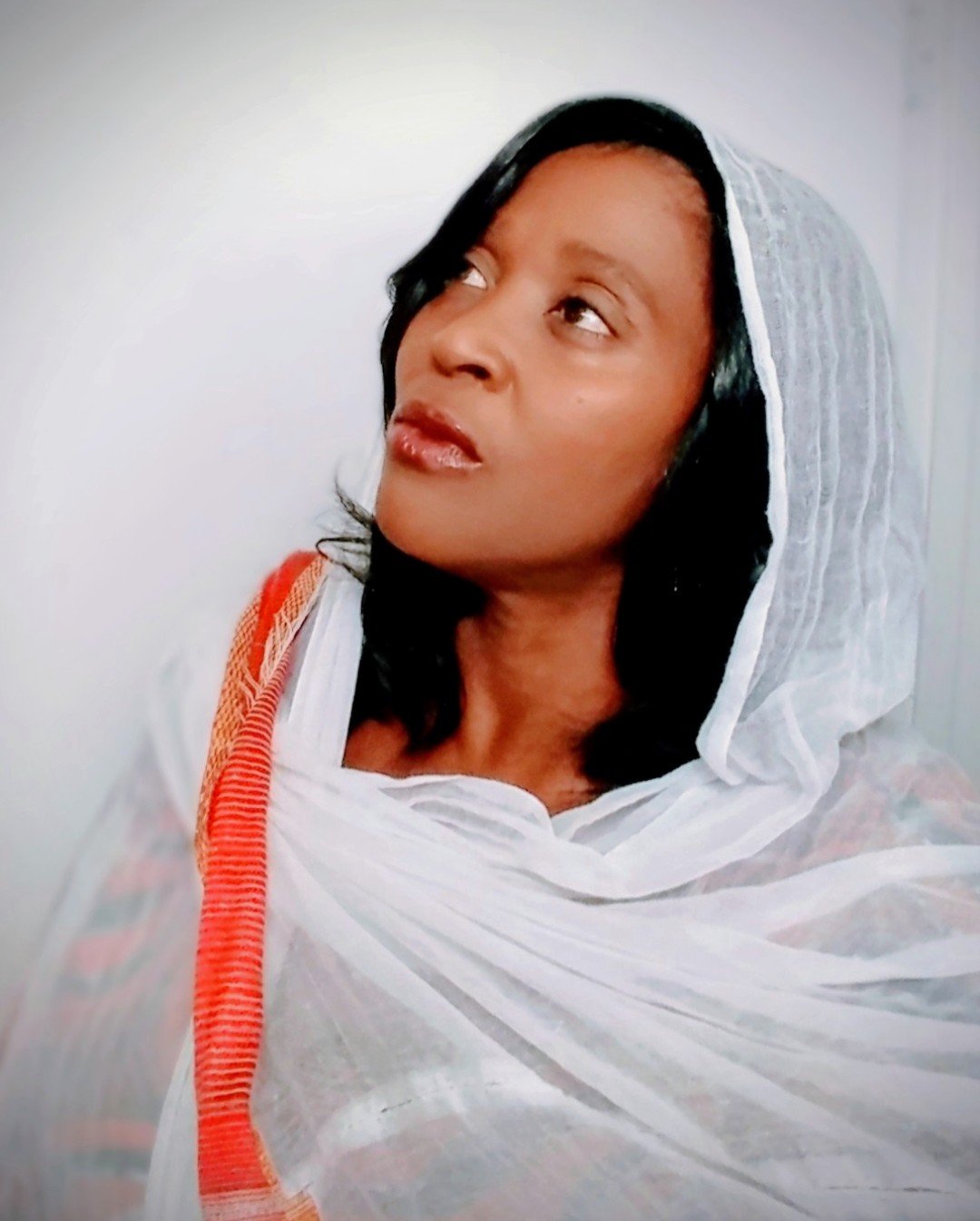 A woman with medium skin tone wearing a white headscarf and attire, gazing upward, with black hair visible.