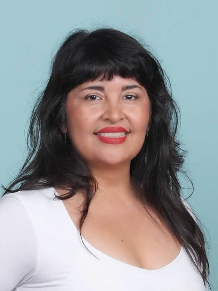 A woman with dark hair, bangs, red lipstick, smiling, wearing a white top, against a light blue background.