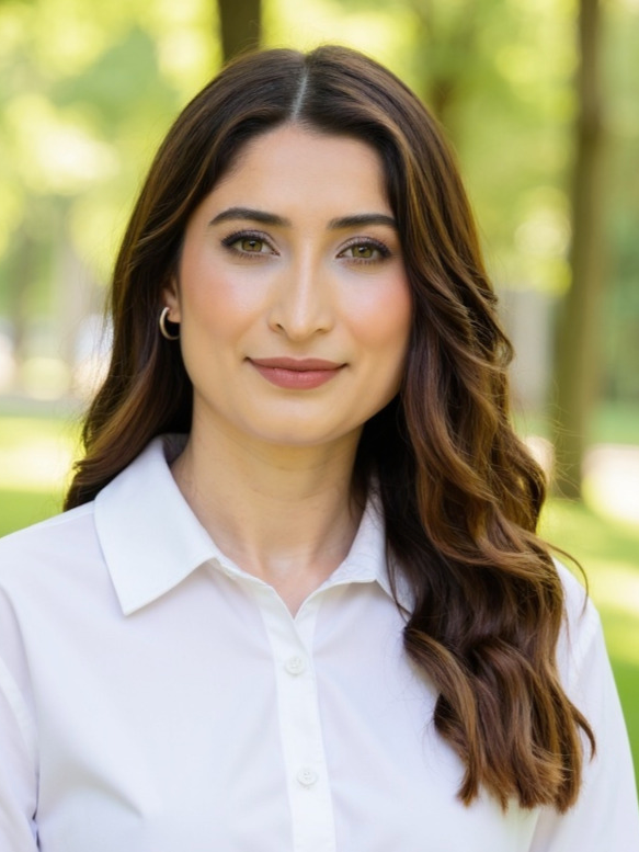 A woman with long wavy brown hair, wearing a white collared shirt, standing outdoors in a park-like setting with green trees in the background.