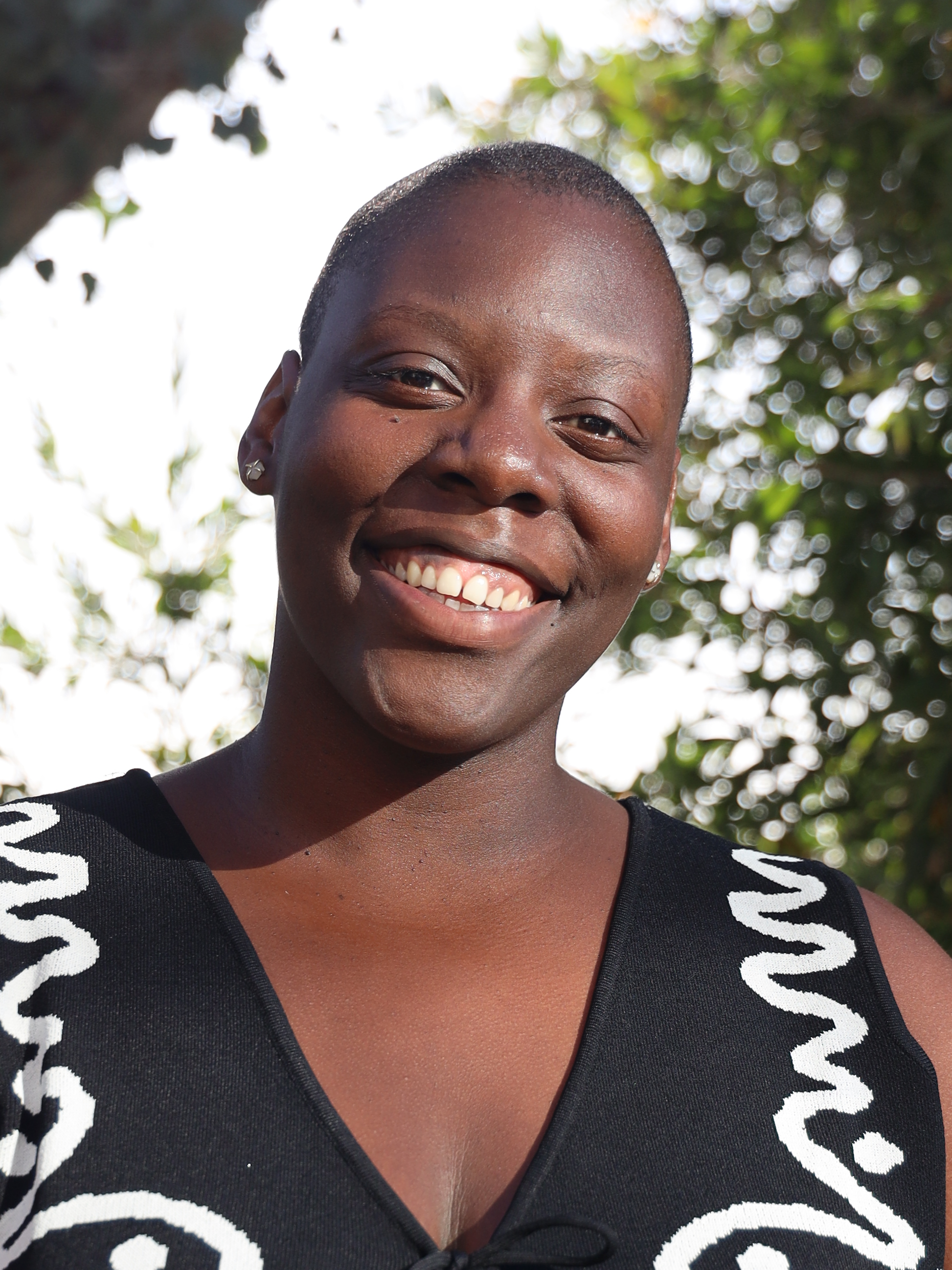 Close-up of a smiling woman with short hair, wearing earrings, outdoors with sunny sky and trees in the background.