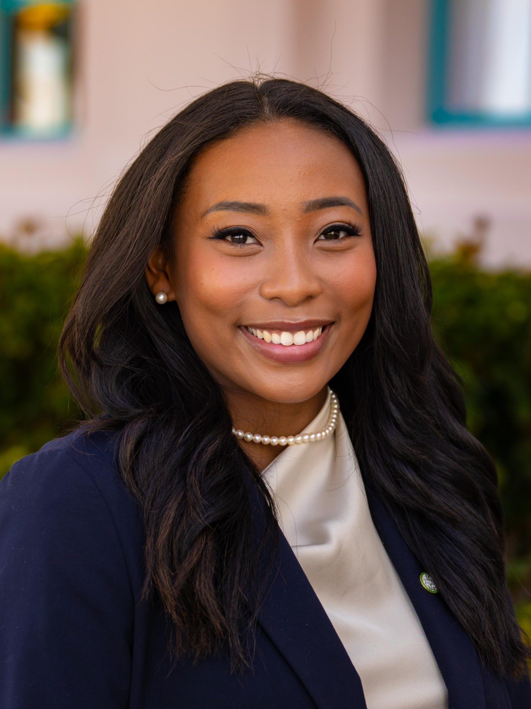 Close-up of a smiling woman with dark hair, pearl earrings, and a pearl necklace, wearing a navy blazer and cream top, outdoors with green shrubbery and colorful background.