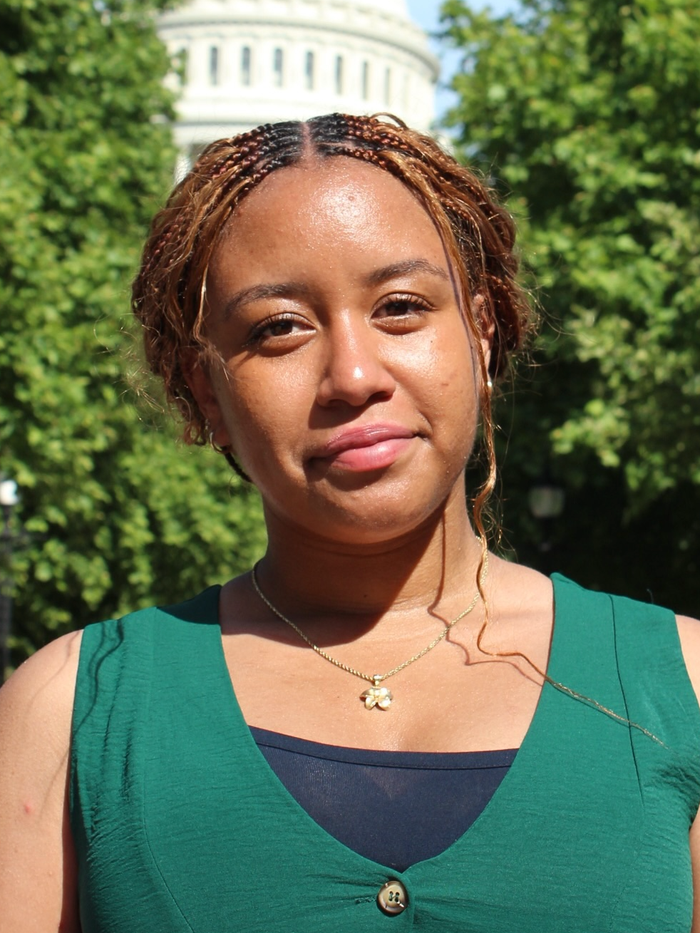 A woman with braided hair outdoors, wearing a green sleeveless top and a gold necklace with a flower pendant, standing in front of green trees and a white dome building.