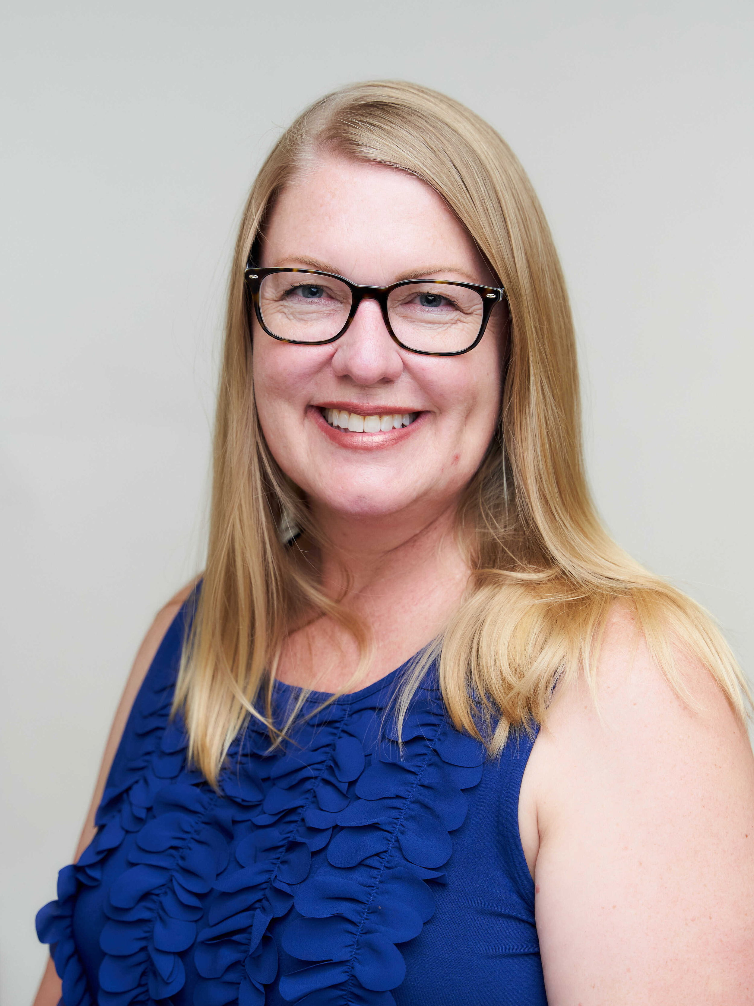 A woman with blonde hair, wearing black glasses and a blue sleeveless dress with ruffled details, smiling in front of a plain light gray background.