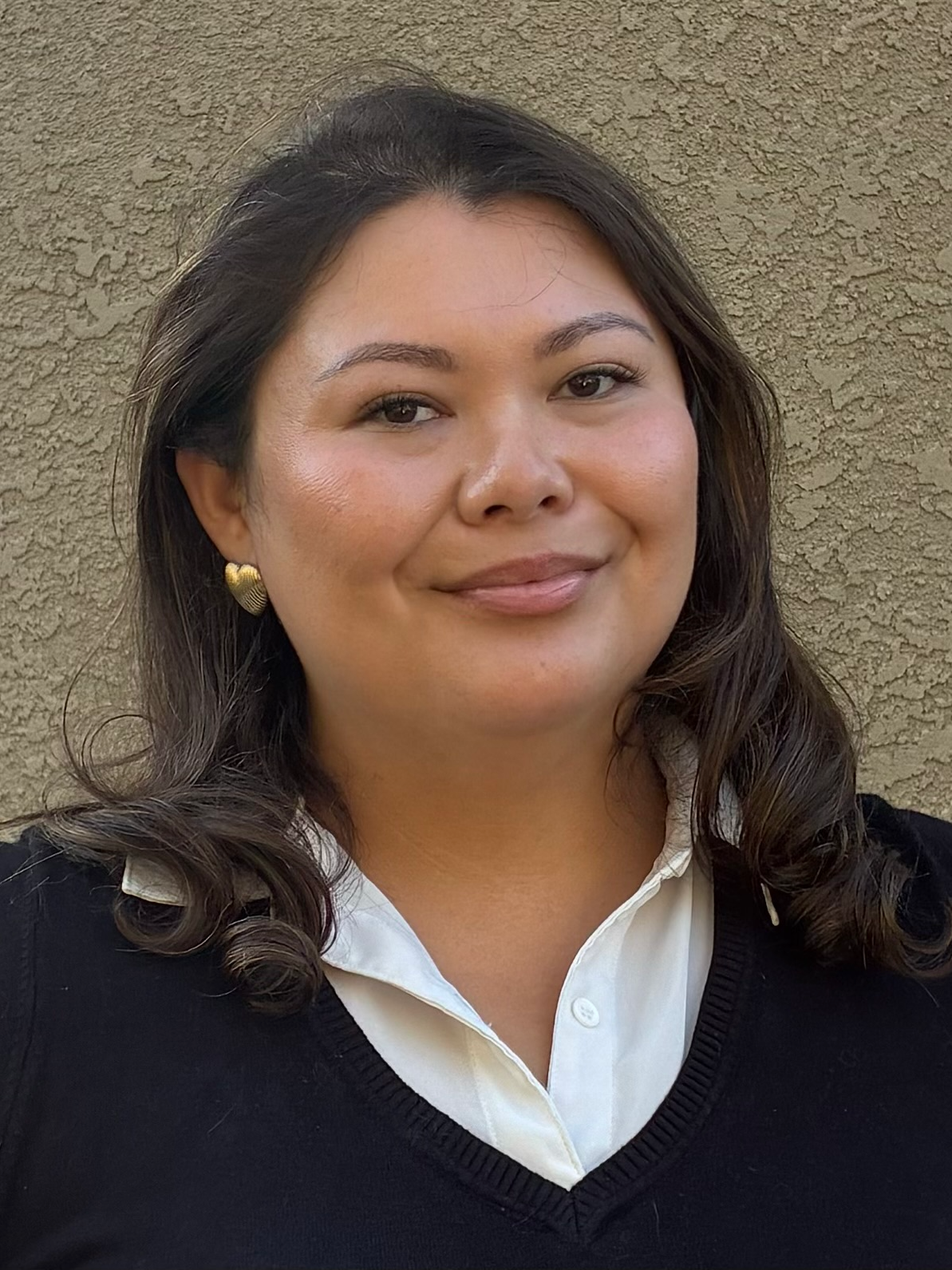A woman with dark hair, wearing heart-shaped earrings, a white collared shirt, and a black sweater, standing against a textured beige wall.