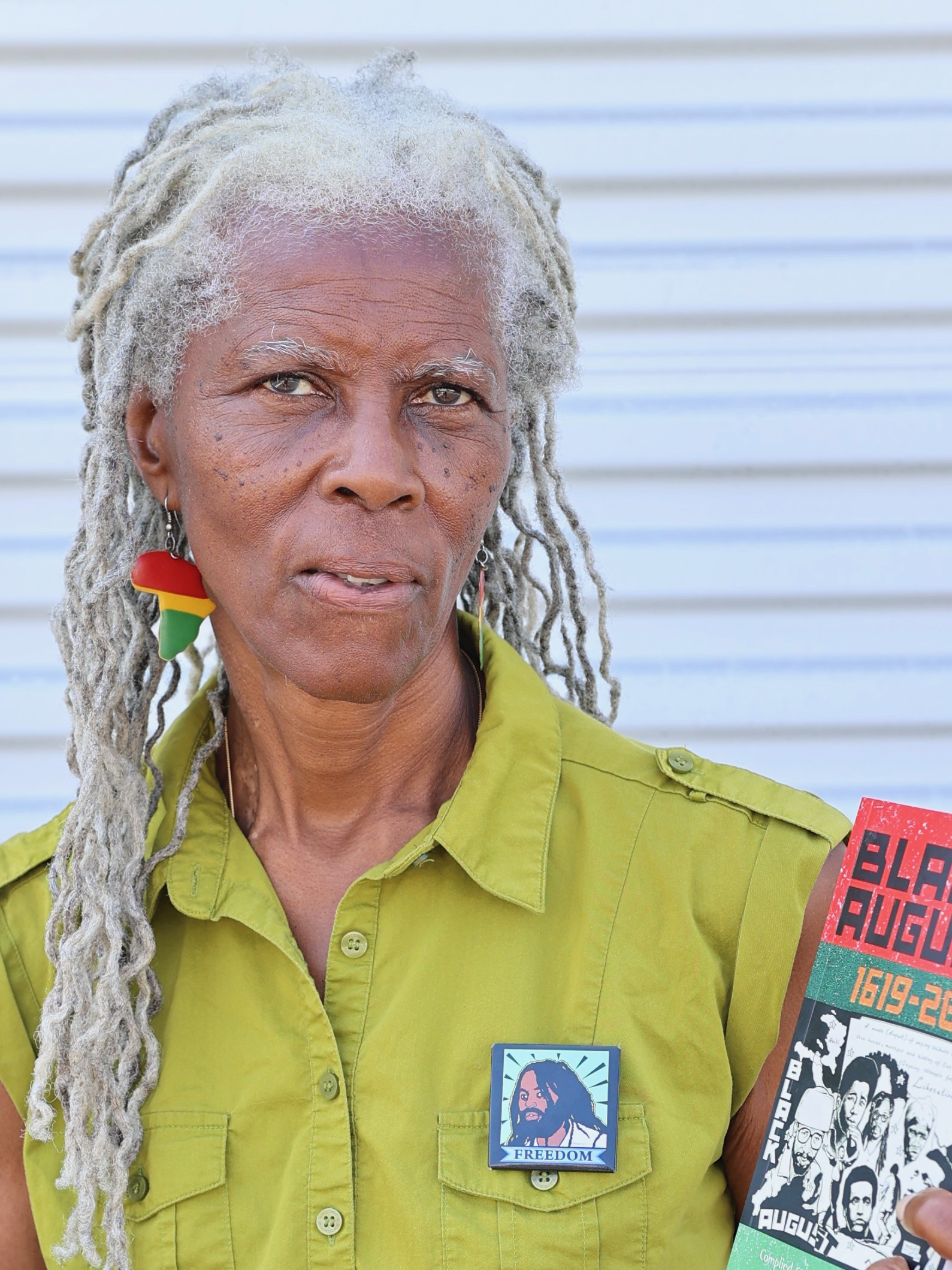 Close-up of an elderly person with gray dreadlocks wearing a green shirt and a badge with a portrait and the word 'FREEDOM' on it, holding a pamphlet or flyer.