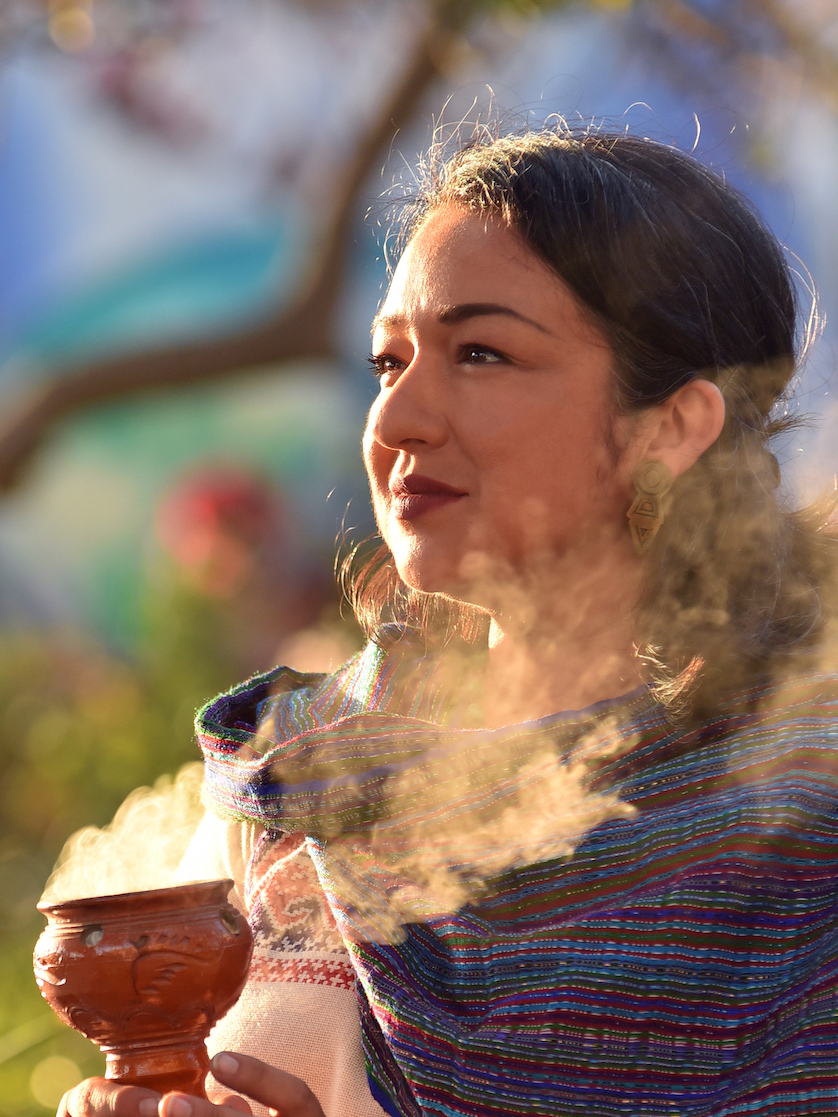 A woman holding a steaming clay pot outdoors on a sunny day, wearing a colorful striped shawl and carrying a traditional earring.
