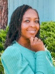 A woman with dark curly hair wearing a turquoise sweater, smiling outdoors with greenery and a blue background.
