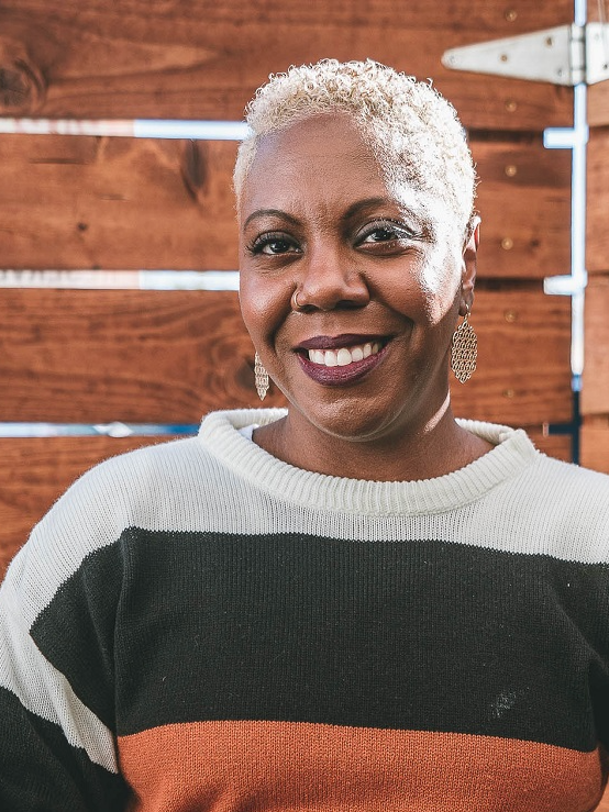 A smiling woman with short gray hair, wearing a black, white, and orange color-block sweater and earrings, standing outdoors in front of a wooden fence.