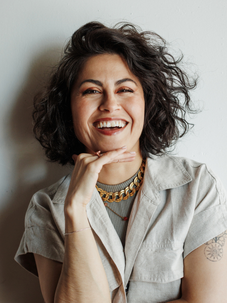 Young woman laughing with short curly hair, wearing a light-colored shirt, gold necklace, and rings, against a plain white background.
